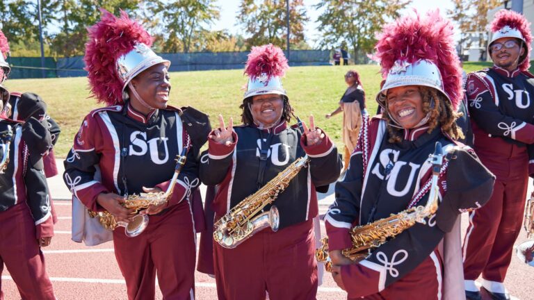 A group of Raleigh marching band members in uniform, holding saxophones, smile and pose for the camera on an outdoor track with trees in the background.
