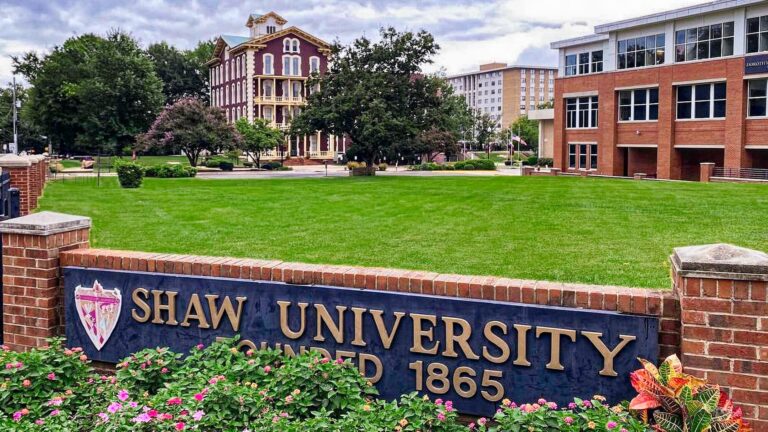 Shaw University campus green space with brick buildings in the background. A sign in the foreground reads, 